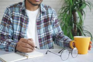 young man writing on notepad while holding a coffee mug photo