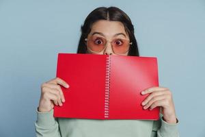Chica adolescente fresca y positiva se cubre la cara con un cuaderno foto