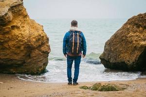 Hombre con una mochila de pie cerca de una roca frente a un hermoso mar foto