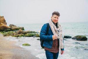 Hombre con una mochila de pie cerca de una roca frente a un hermoso mar foto