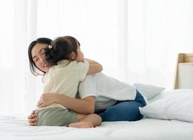 Madre asiática con rostro sonriente abraza a su hija joven en el dormitorio