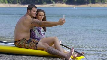 Pareja sentada junto al lago con tablas de paddle surf tomando selfies video