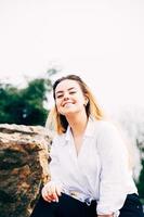 a pretty long haired girl smiling, sitting on a big stone in a park photo