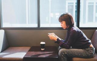 Joven mujer asiática a través de teléfono móvil en el café con una taza de café foto