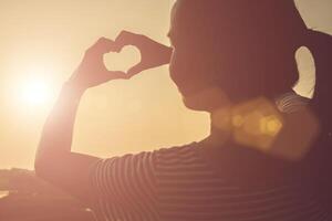 manos de mujer joven en forma de corazón contra la hermosa puesta de sol. foto