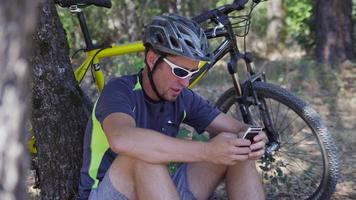 Hombre con bicicleta de montaña se sienta tomando un descanso y revisando el teléfono celular video