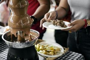 Fuente de chocolate, máquina de catering con brochetas de frutas en rústica mesa de buffet foto