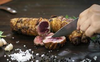 Slicing a serving of organic roast beef roll with knife on wood table with garlic pepper and salt in Melbourne Australia