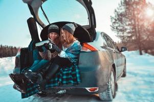 Happy friends in winter forest. Two girls sit in trunk drinking coffee