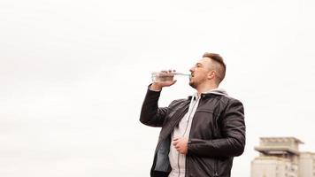 Un hombre con una chaqueta de cuero bebe agua de una botella contra el cielo foto