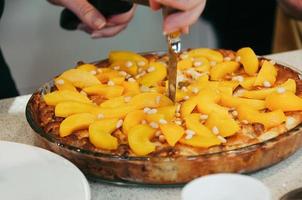 Delicious vegan peach cake decorated with pine nuts, whole, in round glass pan, on table. Selective focus. Colorful healthy juicy. Side view, female hand with knife cutting the cake photo