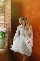 Young blonde woman in white dress dancing by the window, fresh pink flowers bouquet in beautiful vase, bright sienna wall in the backdrop. Romantic, love, happiness concept photo