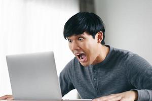 sorprendido hombre asiático mirando la pantalla de la computadora portátil en la sala de estar. foto