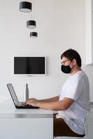 Joven en máscara protectora negra trabajando con un portátil desde casa