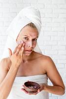 Joven mujer caucásica vistiendo toallas blancas aplicando exfoliante en su rostro foto