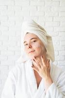 Happy young caucasian woman in white bath towel applying face cream at home photo