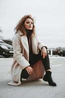 A girl with red curly hair in white coat poses on outdoor parking
