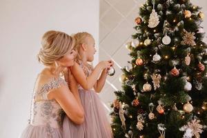 hermosa joven madre sostiene a su hija que decora un árbol de navidad foto