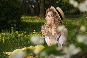 Beautiful young woman with blonde hair in straw hat drinks wine photo