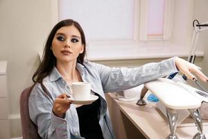 Woman does manicure in a beauty salon and holds a white cup of coffee photo