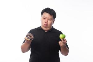 Young funny fat Asian man choosing between chocolate donuts and apple photo