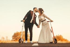 Smiling wedding couple riding a on scooters along the road at sunset photo