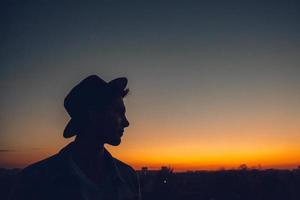 Silueta de un retrato de hombre con sombrero viendo la puesta de sol sobre la ciudad foto