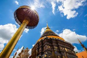 Old ancient golden pagoda with an umbrella of Wat Phrathat Lampang Luang temple with sun star shape on cloudy blue sky, Lampang province, Thailand.
