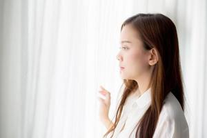 Retrato de una hermosa joven asiática de pie junto a la ventana y sonreír mientras se despierta con el amanecer por la mañana, estilo de vida y concepto de relajación. foto