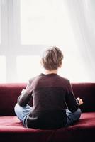 A portrait of a boy sitting on a sofa in a lotus pose, view from his back, against the light window, soft focus photo