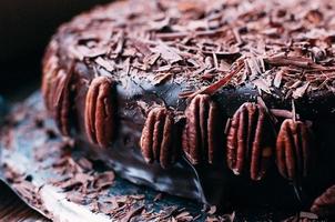 Macro view of whole dark chocolate cake with beautiful icing, chips and pecan nuts on the side on the metal dish. Selective focus. Luxurious glaze. Image for menu or confectionery catalog photo