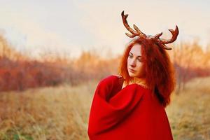 Woman in long red dress with deer horns in autumn forest