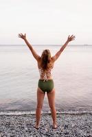 Young woman standing on stony beach with outstretched arms dancing photo