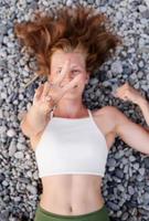 Mujer sonriente joven tumbado en la playa de piedra, cubriendo el rostro con las manos foto