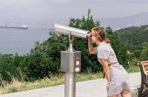 Woman using stationary binoculars against background of sea and city photo
