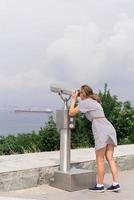 Woman using stationary binoculars against background of sea and city photo