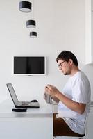 Young smiling man working with laptop from home, drinking water photo