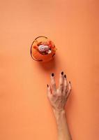 Mano de mujer aterradora con uñas negras tratando de obtener calabaza con dulces, plano sobre fondo naranja foto