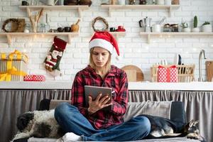 Joven mujer rubia con gorro de Papá Noel trabajando en tableta sentado en el sofá foto