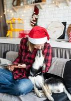 Mujer con gorro de Papá Noel trabajando en tableta sentada en el sofá con su perro