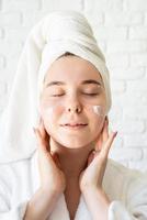 Woman in white bath towels applying face cream doing spa procedures photo