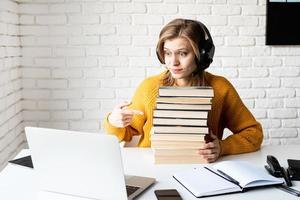Mujer joven en auriculares negros usando laptop sosteniendo una pila de libros