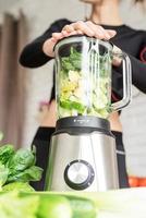 Joven mujer sonriente rubia haciendo batido verde en la cocina de casa foto