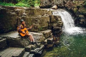 Man playing guitar sitting by a waterfall photo