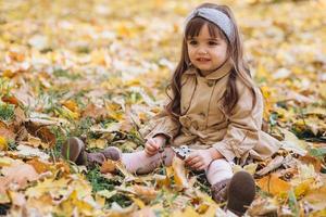 niña con un abrigo beige sentada entre hojas en el parque de otoño.