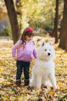 Happy little girl walks with a white Samoyed dog in the autumn park photo