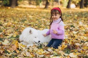 Happy little girl walks with a white Samoyed dog in the autumn park photo