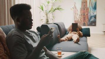 Man sitting using smartphone and girl lying on sofa watching tablet video