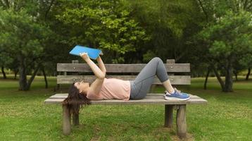Young woman in sportswear lying on the wooden bench reading a book photo