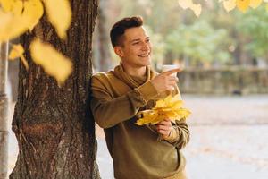 Guy with a bouquet of leaves drinking coffee in autumn park photo